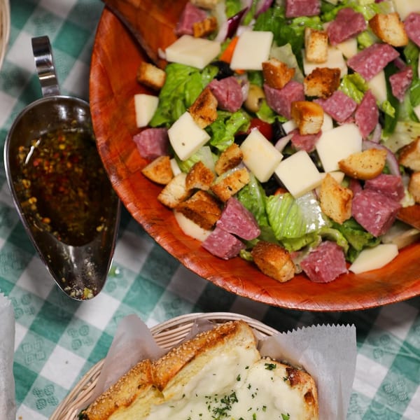 Antipasto salad with salami, cheese, and croutons, alongside garlic bread and dressing on a checkered tablecloth.