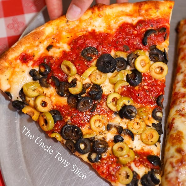 A hand holds a slice of Tony's Brooklyn Pizza with black and green olives on a red and white checkered tablecloth.