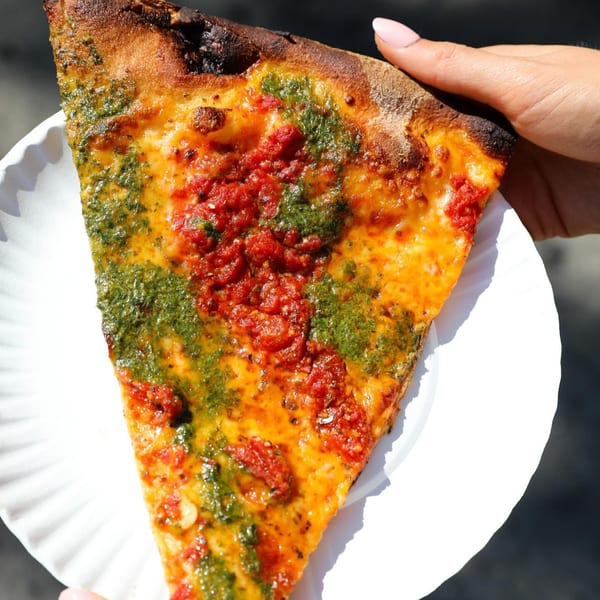 A hand holds a slice of Tony's Brooklyn Pizza with red sauce, pesto, and cheese on a paper plate.